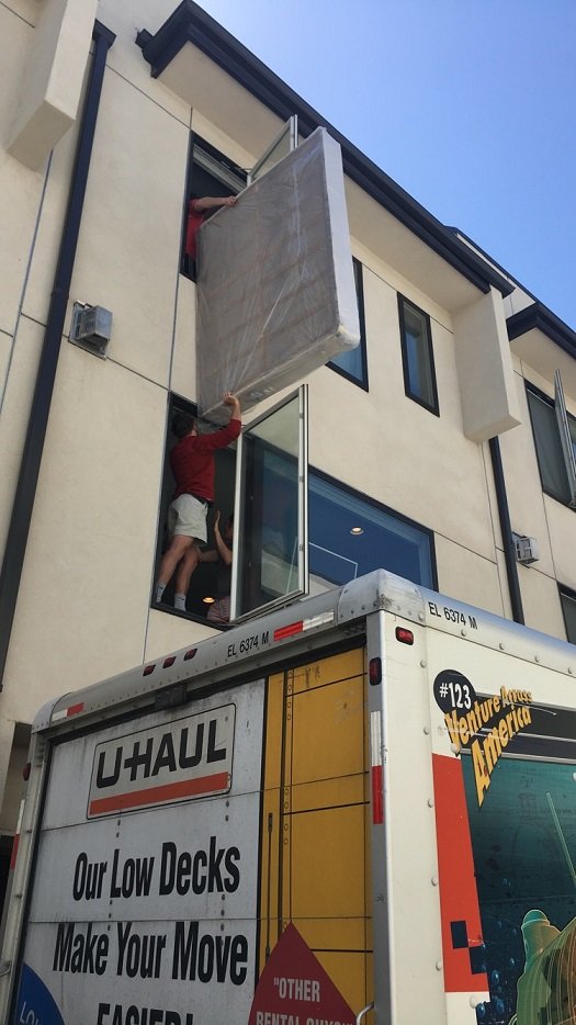 Movers carefully lifting a wrapped mattress through a third-floor window
